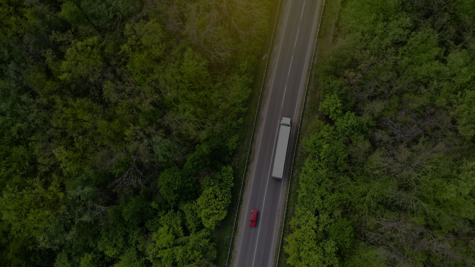 aerial view of a white semi truck and a red car traveling on a two lane highway with green trees on either side